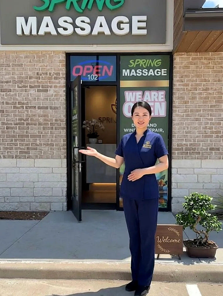 A therapist in navy uniform holding open the door of suite 102, beside a Welcome board and a bonsai on the pavement